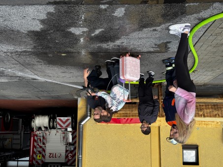 Eine Schülerin hat einen Feuerwehrschlauch in der Hand und löscht ein kleines Feuer. Ein Feuerwehrmann hilft dabei.